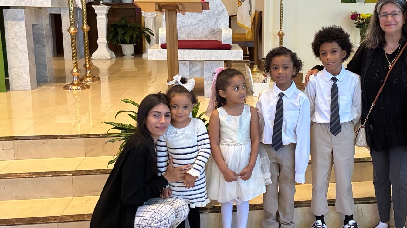 A group of five children and two adults posing together in a church setting. The children are dressed in formal attire, with one girl wearing a white dress and others in suits. The background includes church decor and a pulpit.